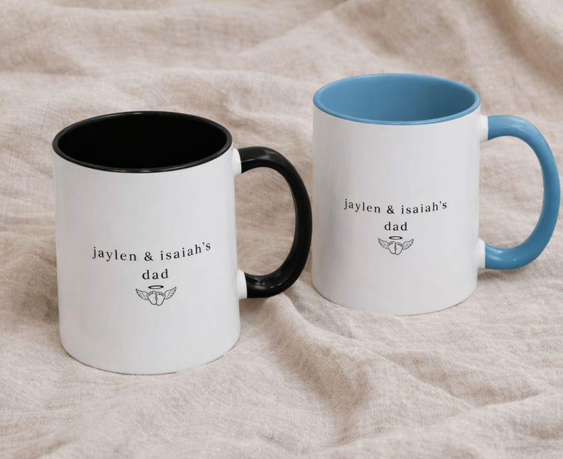 Close-up of custom baby loss dad mug showing “jaylen & isaiah’s dad” text with small angel baby symbol, white ceramic mug with black and blue rim and handle.