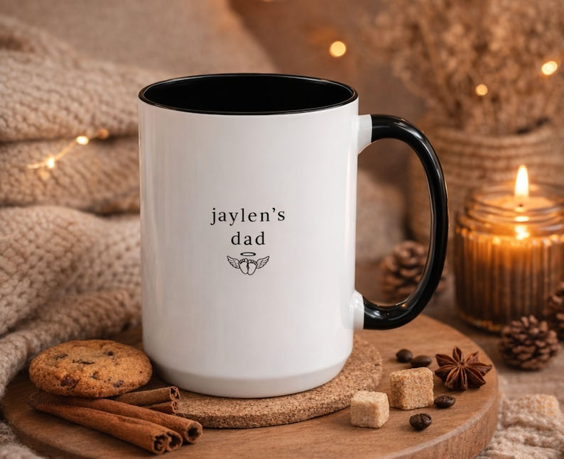 Memorial mug for angel dad, personalized baby names on white ceramic cup with black handle, lifestyle photo with warm soft lighting and minimal decor.