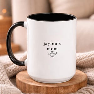White ceramic memorial mug with black handle and interior, personalized ‘jaylen’s mom’ text with small wing symbol, photographed on round wooden coaster with warm, soft living room decor.