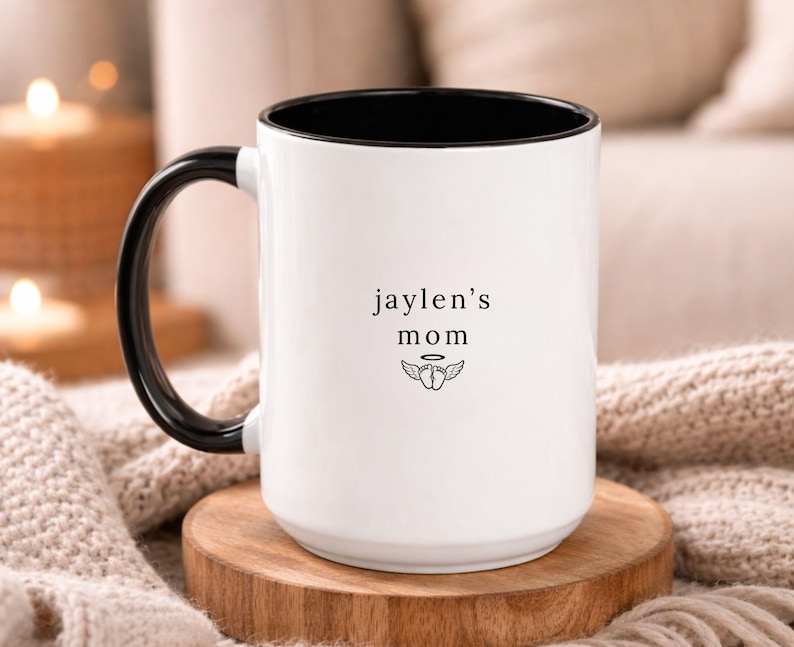 White ceramic memorial mug with black handle and interior, personalized ‘jaylen’s mom’ text with small wing symbol, photographed on round wooden coaster with warm, soft living room decor.