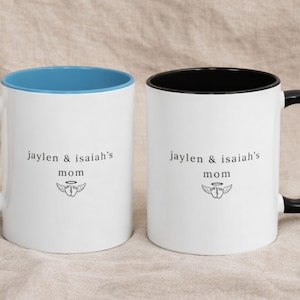 Two personalized remembrance mugs with blue and black handles, white ceramic with colored interiors, custom ‘jaylen & isaiah’s mom’ text and wing icon, photographed on beige fabric for a calm memorial gift style.