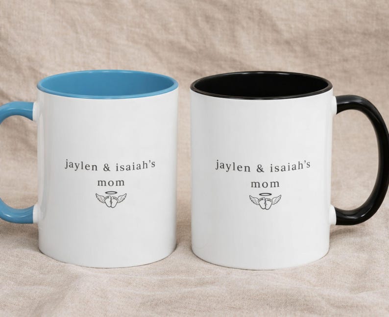 Two personalized remembrance mugs with blue and black handles, white ceramic with colored interiors, custom ‘jaylen & isaiah’s mom’ text and wing icon, photographed on beige fabric for a calm memorial gift style.