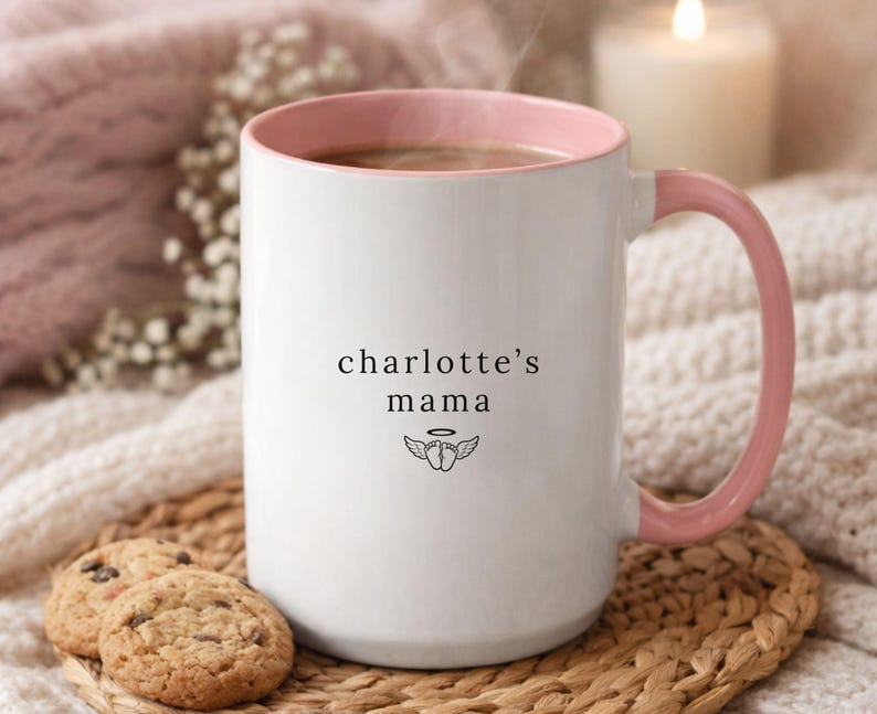 Personalized remembrance mug with pink handle and rim, ‘charlotte’s mama’ text and small angel wing icon, styled on woven coaster with cookies and cozy knit blanket background
