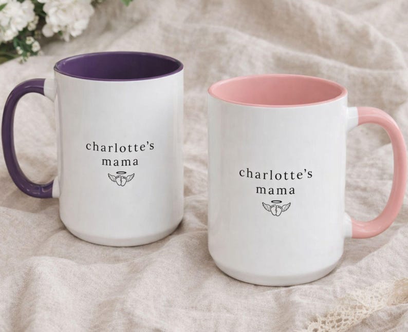 Set of two personalized loss mom mugs, white ceramic with purple and pink handles and colored interiors, ‘charlotte’s mama’ design, resting on neutral linen bedding for a soft comfort gift look.