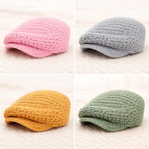May include: Four crochet newsboy caps in pink, gray, mustard yellow, and green. The caps have a classic, rounded shape with a small brim. The hats are made of a textured, knitted material, and are displayed on a light-colored surface.