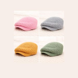 May include: Four crochet newsboy caps in pink, gray, mustard yellow, and green. The caps have a classic, rounded shape with a small brim. The hats are displayed on a light-colored, textured surface.