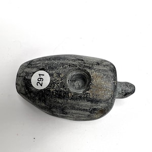 May include: A gray stone pipe with a cylindrical bowl and a small, rounded stem. The pipe has a textured surface and a circular sticker with the number "291" on it.