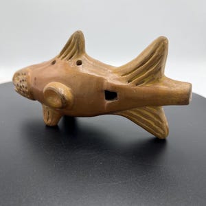 May include: A brown ceramic ocarina shaped like a fish. The instrument has fins, a tail, and a mouth-like opening. It features a series of finger holes and a mouthpiece. The ocarina is a musical instrument.