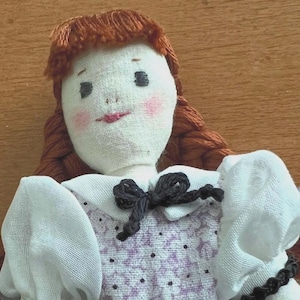 May include: Handmade doll with a fabric face, red yarn hair, and a white dress with a purple floral pattern. The doll has a black bow and a black beaded necklace. The doll's cheeks are blushed pink.