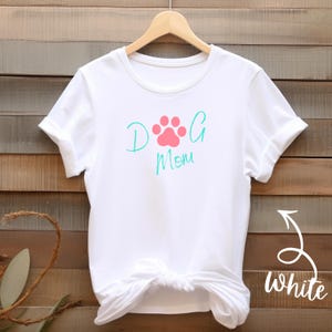 Könnte beinhalten: Weißes T-Shirt mit dem Schriftzug "Dog Mom" in türkisfarbener Schrift und einem rosa Pfotenabdruck-Design. Das Shirt wird vor einem Holzgrund gezeigt.