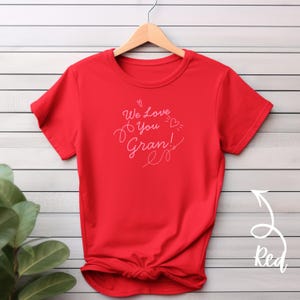May include: A bright red t-shirt with the text "We Love You Gran!" in a cursive font. The design includes a heart and is displayed on a wooden hanger against a white and gray striped background. The shirt is knotted at the bottom.