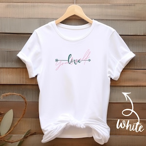 May include: White t-shirt with the words "you love" in pink and green script, with arrows on either side. The shirt is displayed on a wooden background.