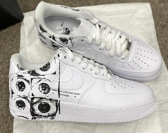 Könnte beinhalten: Weiße Turnschuhe mit schwarzen Augengrafiken an der Seite. Die Schuhe haben weiße Schnürsenkel und Sohlen. Der Text "COMME des GARCONS SHIRT SUPREME" ist auf der Seite des Schuhs aufgedruckt.