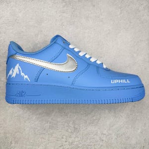 Puede incluir: Zapatilla deportiva azul claro con cordones blancos y un logo plateado. La zapatilla presenta la palabra "UPHILL" y un gráfico de montaña. La suela es azul claro y la palabra "AIR" es visible en el lateral.