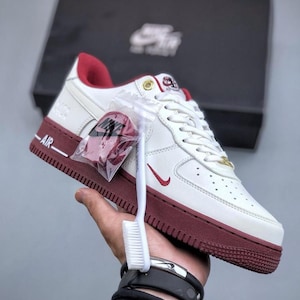 Può includere: Sneakers Air Force 1 bianche e bordeaux con logo swoosh rosso. Le sneakers hanno suola bianca e dettagli bordeaux. Un piccolo accessorio rosso è attaccato ai lacci. Anche una spazzola bianca è attaccata ai lacci.