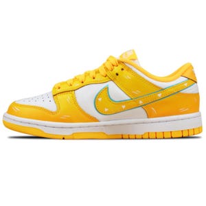 May include: Yellow and white Nike Dunk Low sneakers with a yellow sole and laces. The iconic Nike swoosh is light blue. The shoe has a white toe box with small perforations. The design includes yellow accents and decorative lines.
