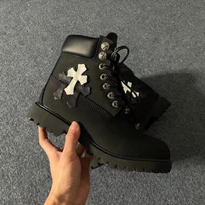 Puede incluir: Botas de cuero negro con un diseño de cruz blanca en el lateral. Las botas tienen suela gruesa, ojales metálicos y un cuello acolchado. Las botas están sujetas por una mano.