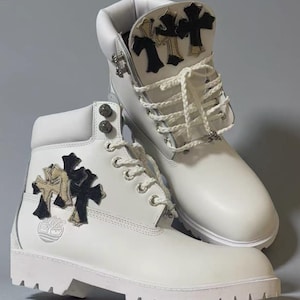Puede incluir: Botas de cuero blanco con aplicaciones decorativas de cruces negras y doradas. Las botas tienen cordones blancos, ojales metálicos y el logotipo de Timberland. Calzado elegante.
