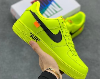 May include: Bright neon green Air Force 1 sneakers with black accents, including the Nike swoosh and a small tag. The word "AIR" is printed on the side of the shoe. A second shoe and a white box are visible in the background.