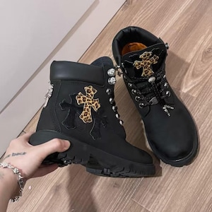 Può includere: Stivali neri con design a croce e dettagli animalier. Gli stivali hanno una chiusura con lacci, dettagli argentati e una suola spessa. Gli stivali sono in pelle e hanno un colletto imbottito.