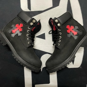 May include: A pair of black leather boots featuring red and grey decorative details. The boots have a lace-up design, silver eyelets, and a chunky sole. The boots are displayed on a black and white patterned surface.