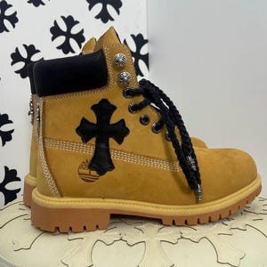 Puede incluir: Bota de trabajo amarillo mostaza con cruz negra y detalles plateados. Cordones negros y cuello acolchado negro. La bota tiene suela marrón y un diseño de cruz negra en el lateral.