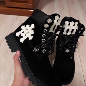Puede incluir: Botines negros con detalles decorativos blancos. Las botas tienen una suela robusta, ojales metálicos y cordones decorativos. La parte superior de las botas es negra con un diseño blanco en el lateral.