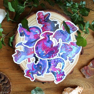 Könnte beinhalten: Eine Sammlung von gestanzten Aufklebern mit verschiedenen Dinosaurier-Illustrationen in einer lebendigen Farbpalette aus Pink, Lila und Blau. Jeder Aufkleber zeigt eine einzigartige Dinosaurierfigur mit stilisierten Mustern.