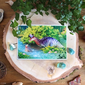 Op de afbeelding: Een rechthoekige kunstdruk met een kleurrijke illustratie van twee hadrosaurus-dinosaurussen in een vijver omringd door weelderig groen gebladerte en roze waterlelies, geplaatst op een grote, ronde houten schijf met schorsrand.