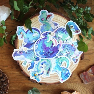 May include: A collection of die-cut vinyl stickers featuring various stylized dinosaur illustrations in shades of blue, teal, and purple. The stickers are arranged in a cluster, showcasing different poses and expressions of these prehistoric creatures.