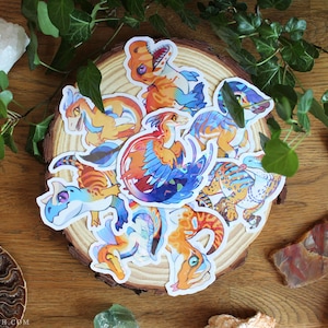 May include: A collection of die-cut vinyl stickers featuring colorful, stylized dinosaur illustrations in shades of orange, blue, and yellow. The stickers are arranged in a cluster on a circular wooden slice, showcasing various poses and expressions.