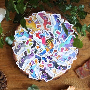 Puede incluir: Una colección de pegatinas troqueladas que representan dinosaurios de estilo caricatura con grandes ojos morados y patrones vibrantes y multicolores. Cada pegatina muestra una paleta de colores única, incluyendo tonos de rojo, azul, amarillo y morado, con bordes blancos.