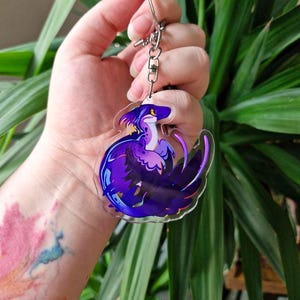 May include: A clear acrylic keyring featuring a stylised illustration of a creature with purple, blue, and yellow feathers. The creature has a long tail, a curved neck, and a yellow eye. It is attached to a silver-coloured metal split ring and chain.