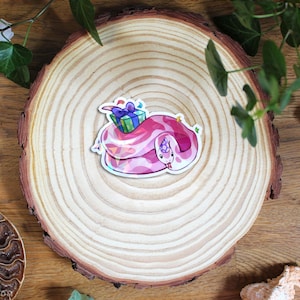Pode incluir: Um autocolante de vinil recortado com uma cobra cor-de-rosa enrolada, usando um chapéu de festa roxo na cabeça e uma pequena caixa de presente verde e azul nas costas. A cobra tem um corpo com padrões rosa e magenta e uma barriga branca.