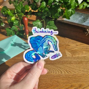 Könnte beinhalten: Ein holografischer Aufkleber mit einem blauen Dinosaurier mit geometrischem Muster, der auf einen kleinen lila Fisch blickt. Über dem Dinosaurier steht der Text thimkning... in dunkelblauer Schrift. Der Aufkleber hat einen weißen Rand und eine reflektierende Oberfläche.