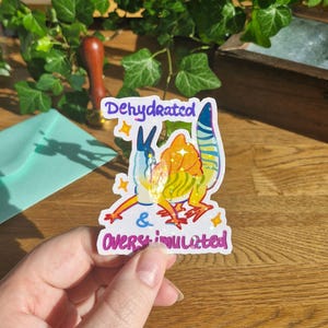 Könnte beinhalten: Ein holografischer Aufkleber mit einer bunten, stilisierten Dinosaurier-Illustration mit gelbem, orangem und blauem Körper. Der Text lautet Dehydrated & Overstimulated in violetter Schrift. Das Design enthält kleine gelbe Sternakzente.