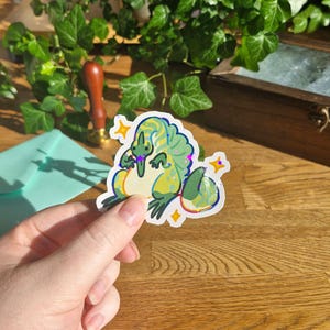 Könnte beinhalten: Ein gestanzter Aufkleber mit einem stilisierten grünen und gelben Dinosaurier mit markanter Rückenflosse, cremefarbenem Bauch und langem Schwanz. Das Design enthält kleine gelbe Sternakzente und eine holografische Oberfläche, die Licht reflektiert.