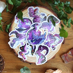 Pode incluir: Uma coleção de adesivos de vinil cortados que apresentam várias ilustrações de dinossauros roxos e azuis com detalhes de penas. Os adesivos estão dispostos em um grupo circular, cada um exibindo uma criatura única com dentes afiados.
