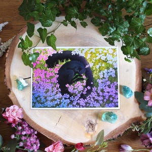 Op de afbeelding: Een rechthoekige kunstdruk met een zwarte kat die in een cirkel is opgerold en slaapt te midden van een levendig veld met kleine roze, paarse, blauwe en gele bloemen. De illustratie gebruikt een verzadigd kleurenpalet om de rustende kat weer te geven.
