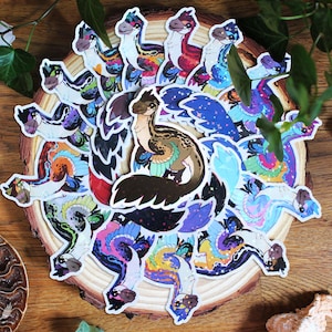 Pode incluir: Uma coleção de autocolantes recortados com dinossauros emplumados ilustrados em várias paletas de cores vibrantes, incluindo azul, roxo, rosa e verde, dispostos num padrão circular sobre uma fatia de madeira redonda.