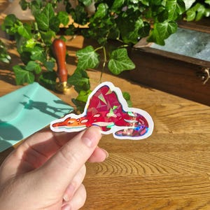 Op de afbeelding: Een gestanste sticker met een gestileerde draak met een geometrisch, veelvlakkig rood en roze patroon. Het ontwerp heeft een holografische afwerking die licht in verschillende kleuren reflecteert, wat een glinsterend effect op het lichaam geeft.