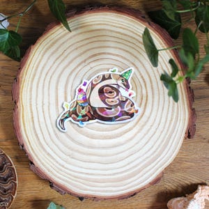 Op de afbeelding: Een gestanste vinylsticker met een bruin en beige gespikkelde slang die een groene feesthoed met stippen draagt. De slang is opgerold rond een kleine, kleurrijke cupcake met een kers en hagelslag, met daaromheen wat confetti verspreid.