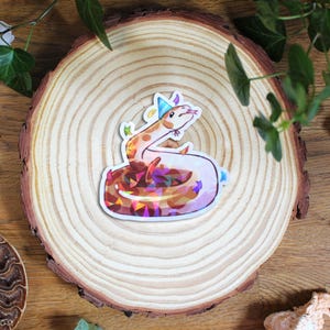 Pode incluir: Um adesivo de vinil cortado com o formato de uma cobra de desenho animado usando um chapéu de festa azul-claro. A cobra tem a parte superior do corpo bege e a parte inferior com um padrão geométrico multicolorido no estilo de vitral.