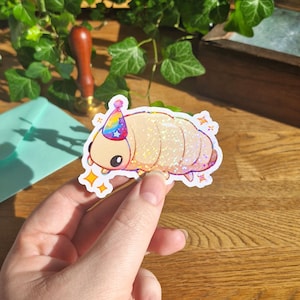 Op de afbeelding: Een gestanste sticker met een lichtbeige, gesegmenteerd pissebedachtig wezen dat een klein, regenboogkleurig feesthoedje met sterren draagt. De sticker heeft een holografische afwerking die licht reflecteert en is versierd met kleine, gele, vierpuntige sterren.