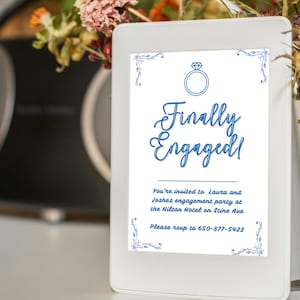 Könnte beinhalten: Eine weiß gerahmte Einladung mit dem Text "Finally Engaged!" in blauer Schrift, mit einer Ringgrafik. Die Einladung wird mit einer Blumenvase und einer Stereoanlage im Hintergrund präsentiert. Die Einladung ist für eine Verlobungsfeier.