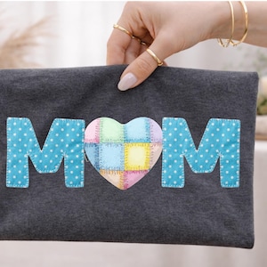 Op de afbeelding: Een donkergrijze vilten etui met het woord "MOM" in blauwe stippenletters en een patchwork hart in het midden. De etui wordt vastgehouden door een hand met gouden ringen.
