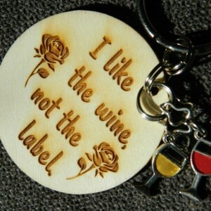 May include: Wooden keychain with a round tag that says "I like the wine, not the label!" with two roses on either side of the text. The keychain has two metal charms attached to it, one with a yellow and black wine glass and the other with a red wine glass.