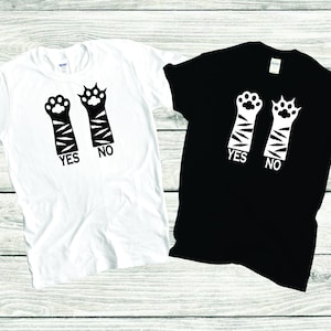 May include: Two t-shirts with a paw print design. One t-shirt is white with black paw prints and the text "YES" and "NO". The other t-shirt is black with white paw prints and the text "YES" and "NO".