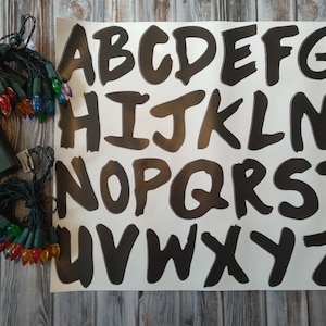 May include: A white sheet of paper with black letters of the alphabet, A through Z, in a handwritten style. Two battery-operated string lights with multi-colored bulbs are on a wooden surface.