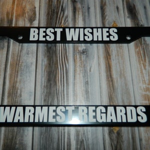 May include: Black license plate frame with white text that reads "BEST WISHES" on the top and "WARMEST REGARDS" on the bottom.
